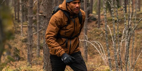 Un homme en veste marron, pantalon noir et bonnet gris se promène dans une forêt dense. Il porte des gants noirs et un sac à dos. La forêt a de grands arbres et des feuilles tombées. Logo « Rukka » en haut.
