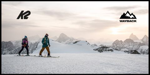 Deux personnes montent une montagne enneigée sous un ciel nuageux, avec des sommets escarpés en arrière-plan. Les logos de K2 et Wayback sont situés dans les coins supérieurs.