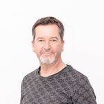 Un homme aux cheveux courts et à la barbe porte une chemise gris foncé à motifs et se tient devant un fond blanc uni. Son expression est calme et il regarde légèrement sur le côté.
