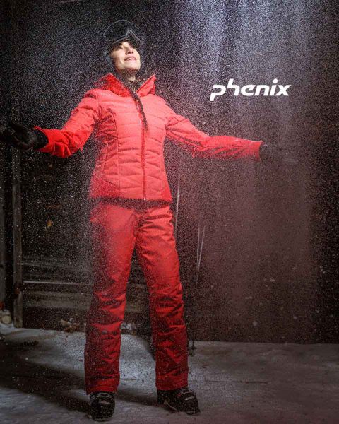 Une personne se tient dehors dans la neige. Elle porte une tenue de ski rouge vif et des gants noirs. Elle a les bras tendus et regarde vers le haut. Au-dessus d'elle, on peut voir le mot « Phenix ». La scène suggère que la personne apprécie le temps hivernal.
