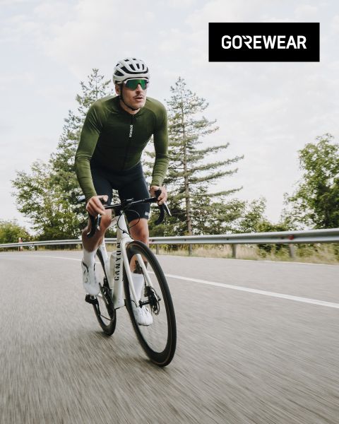 Un cycliste portant une veste verte et un casque roule sur un vélo blanc sur une route. On aperçoit des arbres et un ciel nuageux en arrière-plan. Le logo "GOREWEAR" est visible en haut à droite.