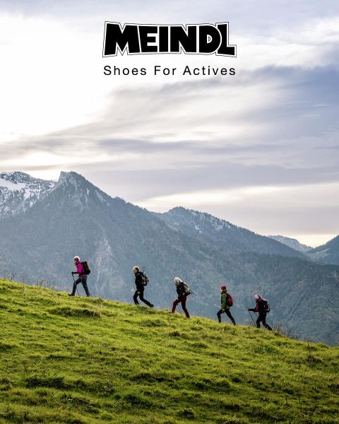 Un groupe de randonneurs avec des sacs à dos monte une colline herbeuse en file indienne. En arrière-plan, on aperçoit des montagnes enneigées et un ciel nuageux. En haut de l'image, on peut lire le texte "MEINDL Shoes For Actives".