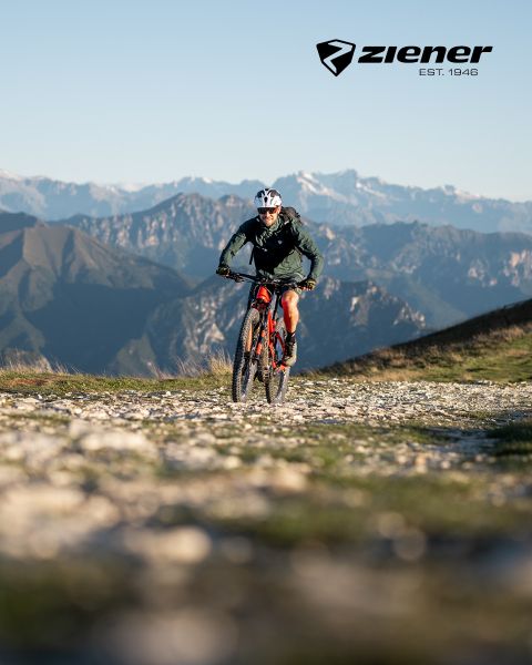 Un cycliste roule sur un chemin de gravier avec son VTT rouge, devant un paysage de montagnes escarpées. Le ciel est dégagé, le cycliste porte un casque et une veste verte. Le logo Ziener se trouve en haut à droite.