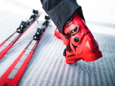 Gros plan d'une personne portant des chaussures de ski rouge vif, debout sur une surface enneigée. À gauche, deux skis rouges sont parallèles l'un à l'autre. L'accent est mis sur la chaussure portant les inscriptions « Redster » et « Atomic », ce qui met en évidence ses détails.