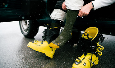 Eine Person, die in einem Fahrzeug sitzt, zieht eine dunkelgrüne Socke an. Daneben liegen gelbe Skischuhe mit schwarzen Riemen auf dem Boden. Schneeflocken fallen herab und die Person trägt eine helle Hose.