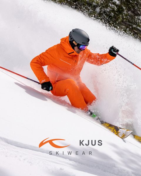 Un skieur en équipement de ski orange vif et casque noir dévale la neige fraîche, soulevant de la poudreuse. L'arrière-plan est enneigé et le logo de KJUS Skiwear apparaît dans la partie inférieure de l'image.