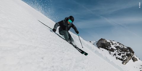 Un skieur en tenue noire et verte dévale une pente enneigée, utilisant des bâtons rouges pour s'équilibrer. En arrière-plan, on aperçoit des rochers enneigés. À gauche, un panneau rouge portant le numéro 11 est visible. Le ciel est clair et bleu.