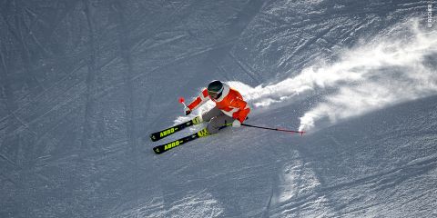 Un skieur en tenue jaune et noire et casque prend un virage serré sur une pente enneigée, projetant de la neige. Le ciel est dégagé et l'équipement du skieur porte le mot « Fischer ».