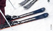 Nahaufnahme eines Paars Skier im Schnee, in dessen Nähe eine Person steht. Die Skier sind dunkel mit hellen Kanten und haben ein sichtbares Markenlogo. Andere Skier sind teilweise im Hintergrund sichtbar.