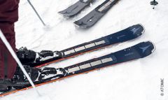 Nahaufnahme eines Paars Skier im Schnee, in dessen Nähe eine Person steht. Die Skier sind dunkel mit hellen Kanten und haben ein sichtbares Markenlogo. Andere Skier sind teilweise im Hintergrund sichtbar.