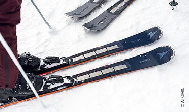Gros plan d'une paire de skis dans la neige, avec une personne debout à proximité. Les skis sont foncés avec des bords clairs et un logo de marque visible. D'autres skis sont partiellement visibles en arrière-plan.