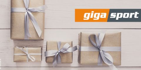Auf einer weißen Holzfläche sind vier goldene Geschenkboxen mit silbernen Bändern angeordnet. In der oberen rechten Ecke ist das Logo von „giga sport“ zu sehen.