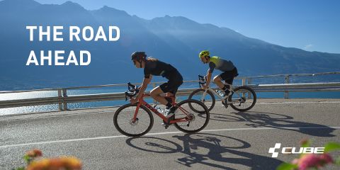 Deux cyclistes roulent sur des vélos de course sur une route de montagne sinueuse avec vue sur un lac et des montagnes pittoresques. Ils portent tous deux des casques et des vêtements de cyclisme. La lumière du soleil projette des ombres. Le texte sur l'image est "LA ROUTE DEVANT" avec le logo Cube dans le coin inférieur droit.