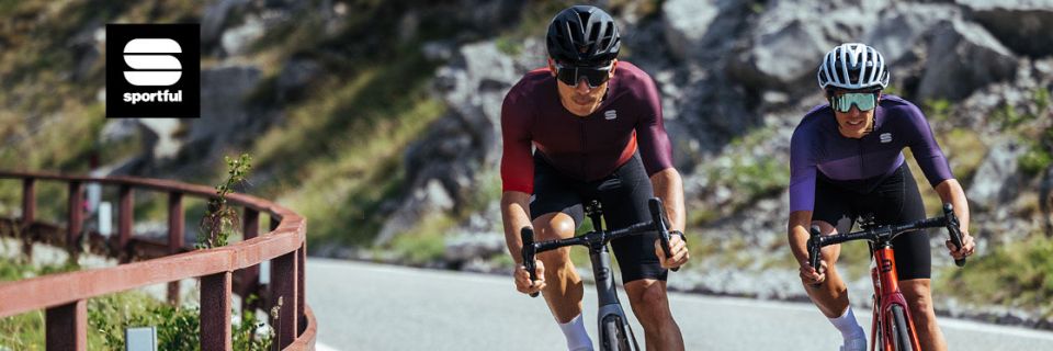 Deux cyclistes en équipement Sportful assorti montent une route de montagne. L'un porte un casque foncé, l'autre un casque blanc. La route est sinueuse, avec une pente rocheuse en arrière-plan. Le logo "Sportful" est visible dans le coin supérieur gauche.