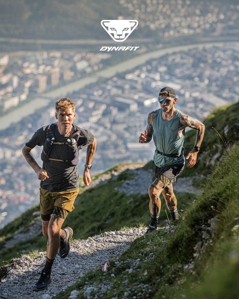 Deux personnes courent sur un sentier de montagne avec une ville et une rivière en arrière-plan. L'une porte un t-shirt noir et un short, l'autre un débardeur bleu et un short. Le logo Dynafit se trouve en haut de l'image.