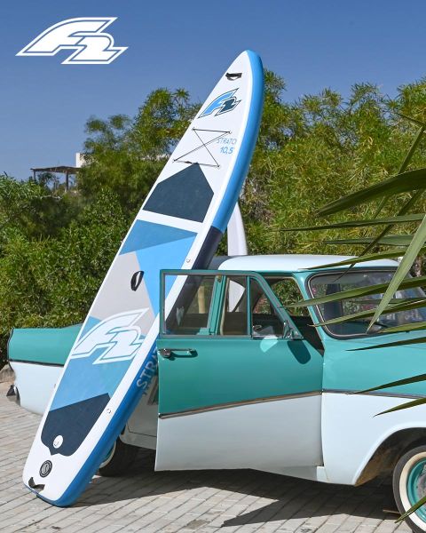 Une voiture ancienne turquoise et blanche est garée, avec une planche de paddle bleu et blanc appuyée contre elle. À l'arrière-plan, on aperçoit des arbres verts, et un logo se trouve dans le coin supérieur gauche.