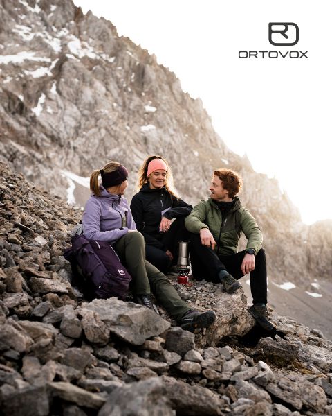 Trois personnes en équipement de plein air sont assises, souriantes et discutant sur un terrain rocheux dans une région montagneuse. Le soleil se couche derrière les sommets. Un sac à dos et une thermos sont posés à proximité. Le logo d'Ortovox se trouve en haut à droite.