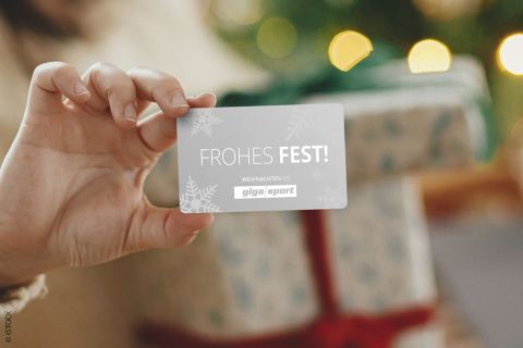 Une personne tient une carte avec l'inscription « Joyeuses Fêtes ! ». À l'arrière-plan, un cadeau emballé avec un ruban rouge repose devant un sapin de Noël décoré de lumières scintillantes.