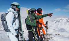 Drei Skifahrer mit Helmen und Skibrillen stehen mit Skiern in den Händen auf einem schneebedeckten Berggipfel. Sie tragen blaue, grüne und orangefarbene Jacken. Einer zeigt in die Ferne. Alle blicken auf die malerische Schneelandschaft unter einem klaren blauen Himmel.