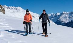 Zwei Personen in Winterkleidung laufen mit Schneeschuhen durch eine verschneite Berglandschaft unter klarem blauen Himmel. Schneebedeckte Gipfel prägen den Hintergrund. Die Person links trägt eine orangefarbene Jacke, die Person rechts eine graue.