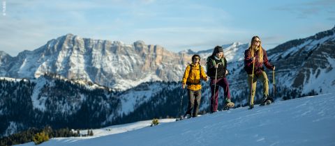 Trois personnes montent une colline enneigée en raquettes, avec des montagnes enneigées en arrière-plan. Ils portent des vêtements d'hiver et des lunettes de soleil et profitent de cette journée ensoleillée dans un paysage enneigé pittoresque.