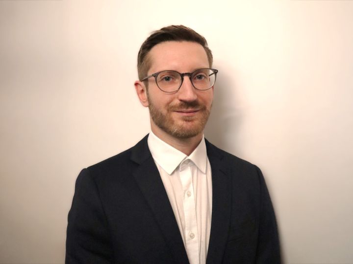 Une personne portant des lunettes, une chemise blanche et un blazer foncé se tient devant un fond clair et uni.