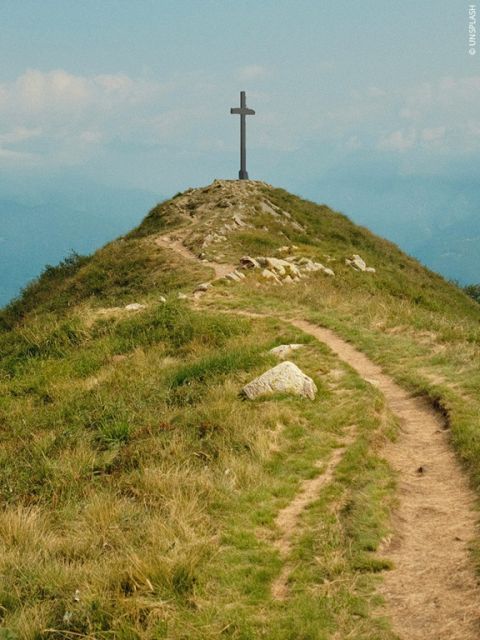 Une colline herbeuse avec un chemin de terre menant à une grande croix au sommet. À l'arrière-plan, le ciel est partiellement nuageux.