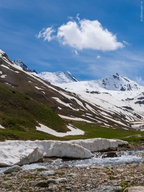 Un paysage de montagne pittoresque avec des sommets enneigés sous un ciel bleu clair. Un petit ruisseau coule au premier plan, bordé de champs de neige et d'une pente herbeuse. Un nuage duveteux solitaire flotte au-dessus.