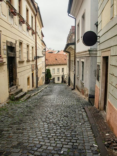 Étroite rue pavée dans une ville européenne, bordée de vieux bâtiments aux murs délavés et aux volets. La rue descend, créant une atmosphère pittoresque et historique sous un ciel nuageux.