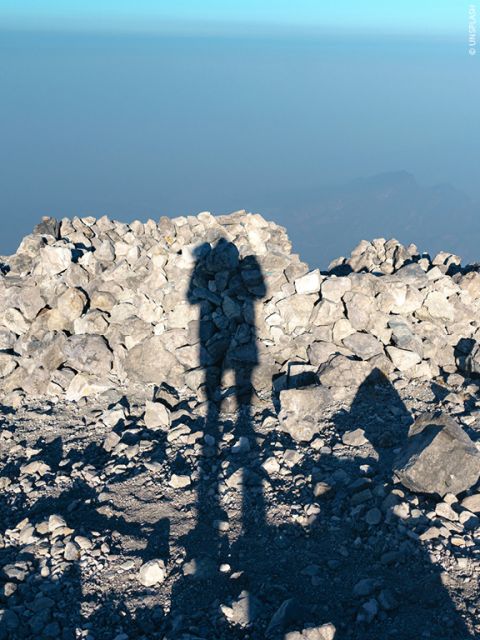 Ombre d'une personne avec un appareil photo sur un terrain rocheux, avec des pierres éparses en arrière-plan et un horizon brumeux et lointain sous un ciel bleu clair.