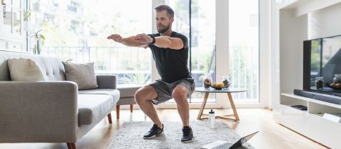 Un homme fait des squats dans un salon avec un canapé gris et une table basse. Il porte une chemise noire et un short gris. Un ordinateur portable est posé sur le sol. À l'arrière-plan, on aperçoit une grande fenêtre donnant sur le balcon.