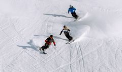 Drei Personen fahren einen schneebedeckten Hang hinunter. Sie tragen Winterkleidung in den Farben Rot, Blau und Schwarz und hinterlassen geschwungene Spuren. Die Sonne wirft Schatten auf den unberührten Schnee und unterstreicht ihre Bewegungen.