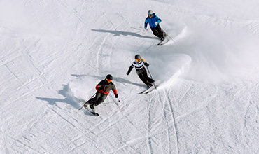 Trois personnes dévalent une pente enneigée. Elles portent des vêtements d'hiver de couleurs rouge, bleue et noire, et laissent des traces sinueuses derrière elles. Le soleil projette des ombres sur la neige immaculée, soulignant leurs mouvements.
