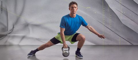 Une personne en t-shirt bleu et short vert fait une fente latérale tout en tenant une kettlebell. En arrière-plan, on aperçoit un mur gris texturé avec des motifs et des lignes discrets.