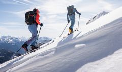 Zwei Personen fahren unter strahlend blauem Himmel einen schneebedeckten Berg hinauf. Sie sind in Winterkleidung mit Rucksäcken und Skistöcken gekleidet und haben einen Panoramablick auf die fernen schneebedeckten Gipfel.
