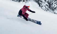 Ein Skifahrer in rot-schwarzer Kleidung und weißem Helm fährt einen schneebedeckten Hang hinunter. Er nutzt Stöcke, um das Gleichgewicht zu halten. Im Hintergrund sind schneebedeckte Bäume zu sehen.