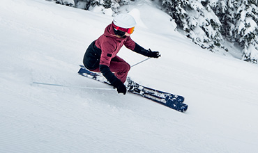 Un skieur en tenue rouge et noire et casque blanc dévale une pente enneigée. Il utilise des bâtons pour garder l'équilibre. Des arbres enneigés sont visibles en arrière-plan.