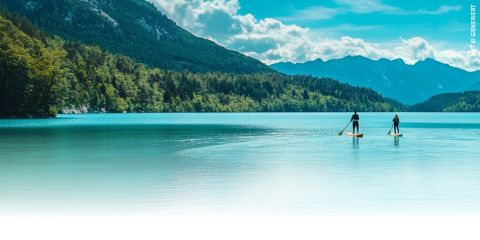 Deux personnes font du stand-up paddle sur un lac turquoise calme, entouré de collines boisées et de montagnes au loin sous un ciel bleu éclatant avec quelques nuages épars.
