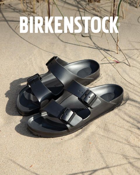 Une personne portant des sandales Birkenstock beiges se tient à côté d'un jeu d'eau dans un paysage de jardin luxuriant. La zone est entourée de verdure et le mot « BIRKENSTOCK » est clairement visible au-dessus de l'image.