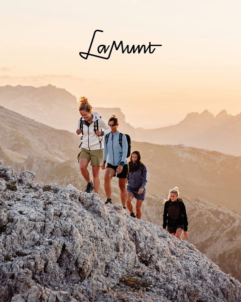 Trois personnes en vêtements d'hiver font du ski dans un paysage montagneux enneigé sous un ciel clair. Le texte est : "LaMunt MOUNTAIN ME-TIME". On aperçoit des sommets enneigés en arrière-plan.