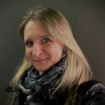 Une femme aux longs cheveux blonds sourit légèrement à la caméra. Elle porte une écharpe à motifs noirs et une veste noire devant un fond sombre et uni.