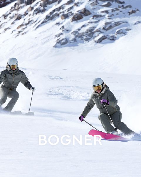 Deux skieurs en équipement coloré. À gauche : personne en jaune avec des lunettes de protection, tirant un traîneau noir. À droite : deux personnes en vestes jaunes et bleues portant des skis. Tous portent des casques et des lunettes de protection. Arrière-plan enneigé avec « BOGNER » dans le texte.