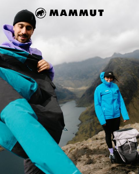 Deux randonneurs en équipement de plein air traversent un terrain rocailleux sous un ciel nuageux, avec une grande montagne en arrière-plan. Ils portent tous les deux des vêtements Mammut, et le logo de la marque est visible en haut de l'image.