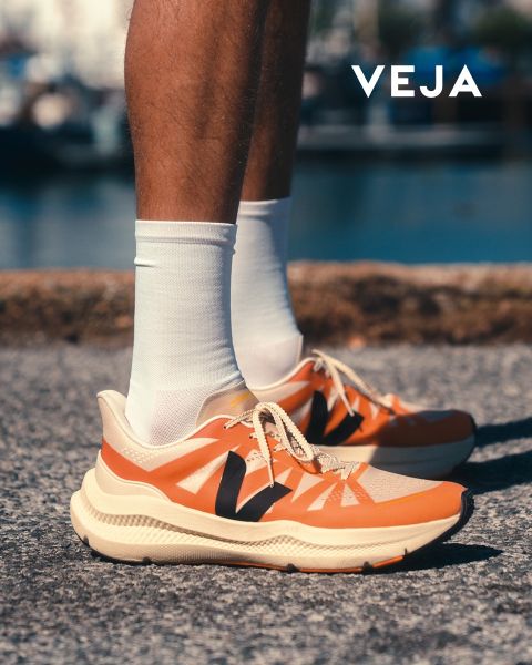 Une personne porte des baskets Veja blanches avec des détails bleu clair et une semelle en caoutchouc. Les chaussures sont associées à un pantalon blanc. L'arrière-plan présente un motif géométrique sur un mur carrelé. Le logo Veja est visible dans le coin inférieur droit.