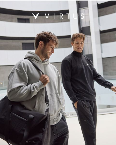 Deux hommes en tenues de sport Virtus, l'un portant un sweat à capuche gris clair avec un sac de sport noir, l'autre une veste noire, sourient et se promènent ensemble dans un hall moderne. Le logo Virtus est visible au-dessus d'eux.
