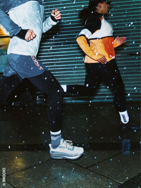 Deux personnes courant de nuit en tenue de sport, avec des gouttes de pluie visibles dans l'air et un trottoir mouillé sous leurs pieds. Les deux coureurs portent des vestes et des chaussures de course et passent rapidement devant un portail métallique.