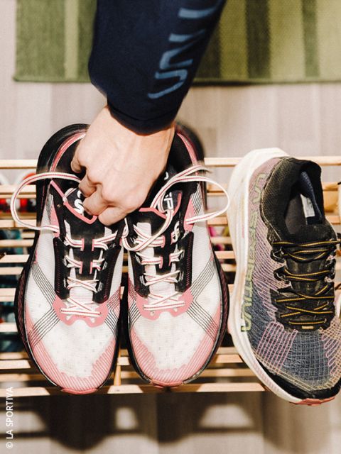 Une personne tient une paire de chaussures de course blanc rosé par les lacets au-dessus d'une étagère à chaussures en bois, à côté d'une seule chaussure de course noire et grise.