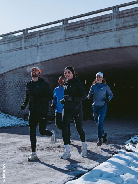 Quatre personnes en tenue de course d'hiver font du jogging ensemble sur un chemin enneigé sous un pont en béton, sourient et profitent de l'exercice en plein air.