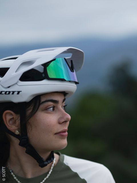 Une jeune femme portant un casque de vélo blanc et des lunettes de soleil réfléchissantes sur la tête regarde sur le côté. L'arrière-plan est flou avec une verdure naturelle et un ciel nuageux et doux.