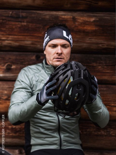Un homme en veste verte et gants examine un casque de vélo noir et est assis devant un mur en bois. Il porte un bandeau noir avec une inscription blanche.